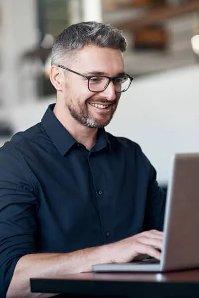 A man with glasses typing on his laptop while smiling